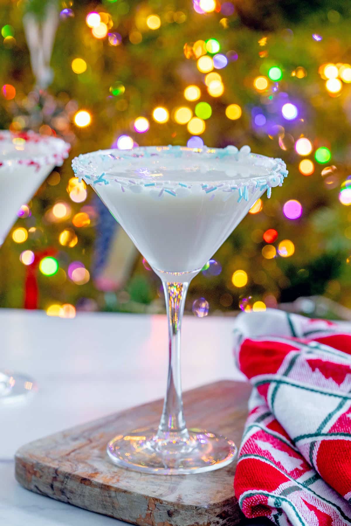 Head-on closeup view of a sugar cookie martini on a wooden board in front of a Christmas tree with lights.