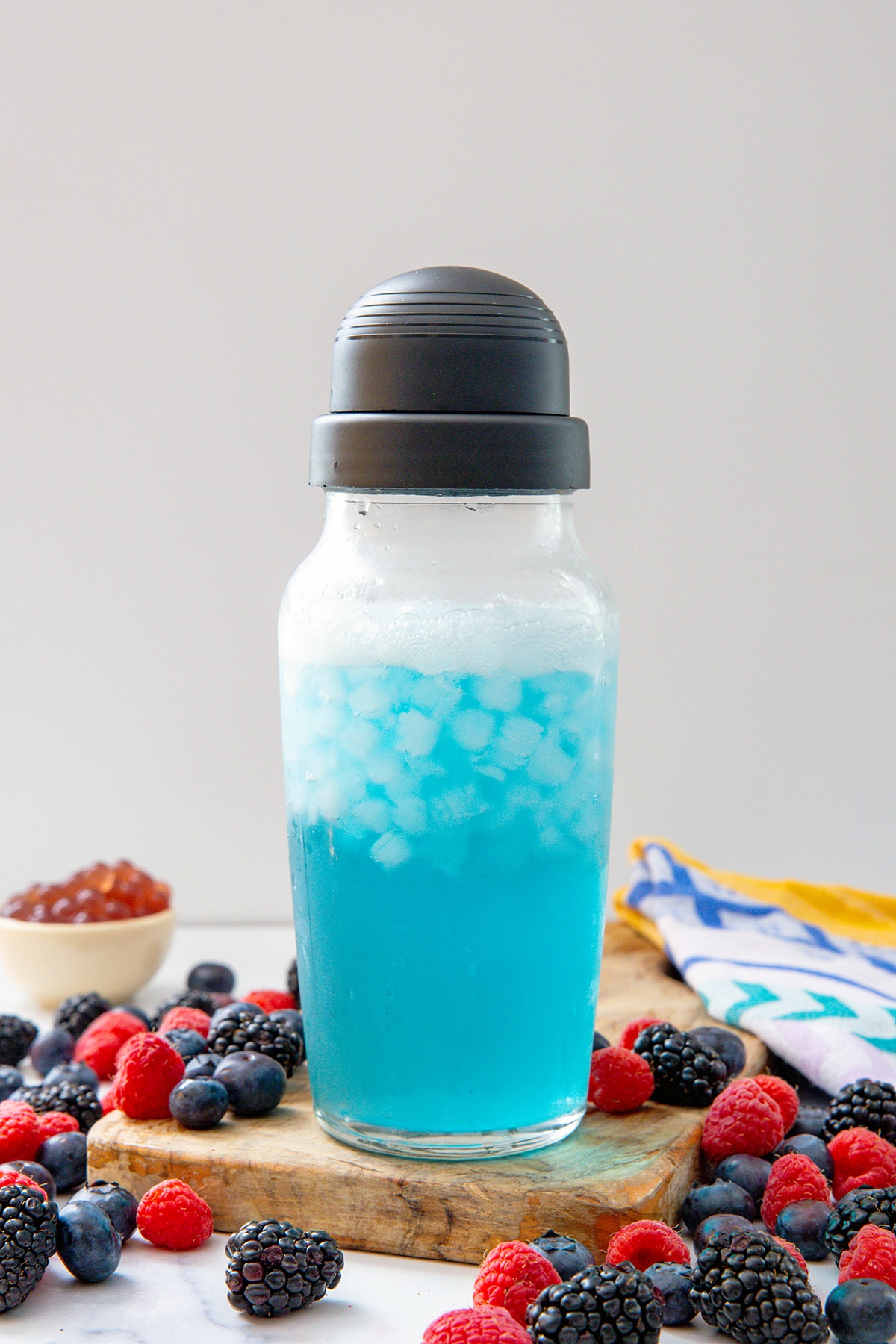 Blue refresher base mixed with water and ice in clear cocktail shaker with berries all around.