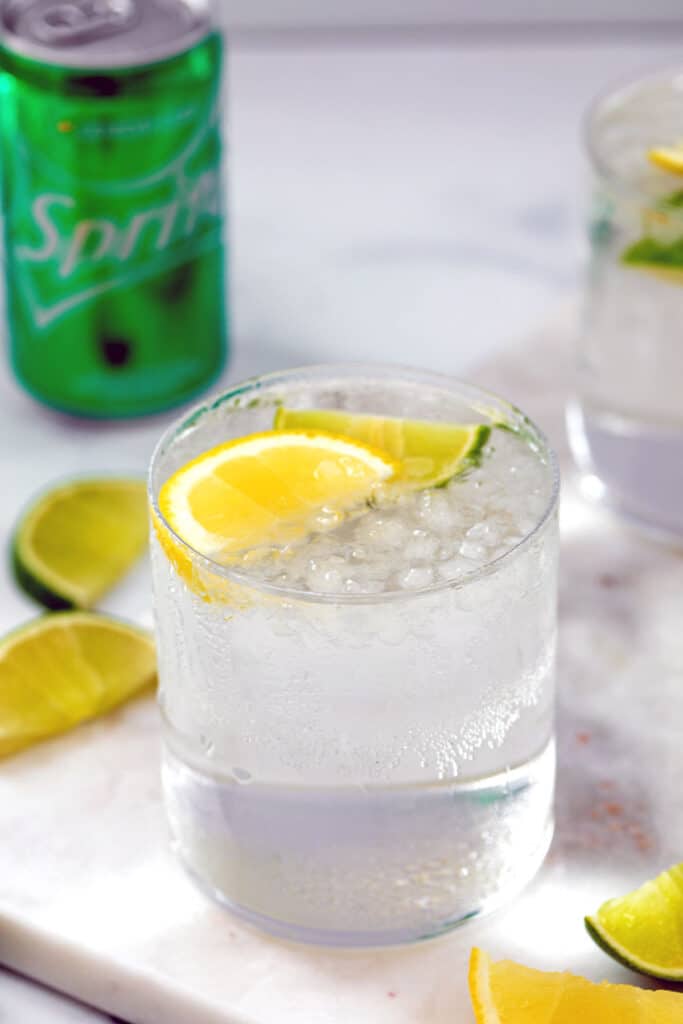 Glass of tequila and sprite with lemon and lime wedges and Sprite can in background..