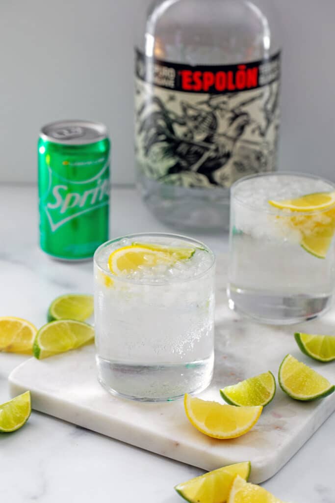 Two glasses of tequila and Sprite with lemon and lime wedges all around and a can of Sprite and bottle of tequila in background.