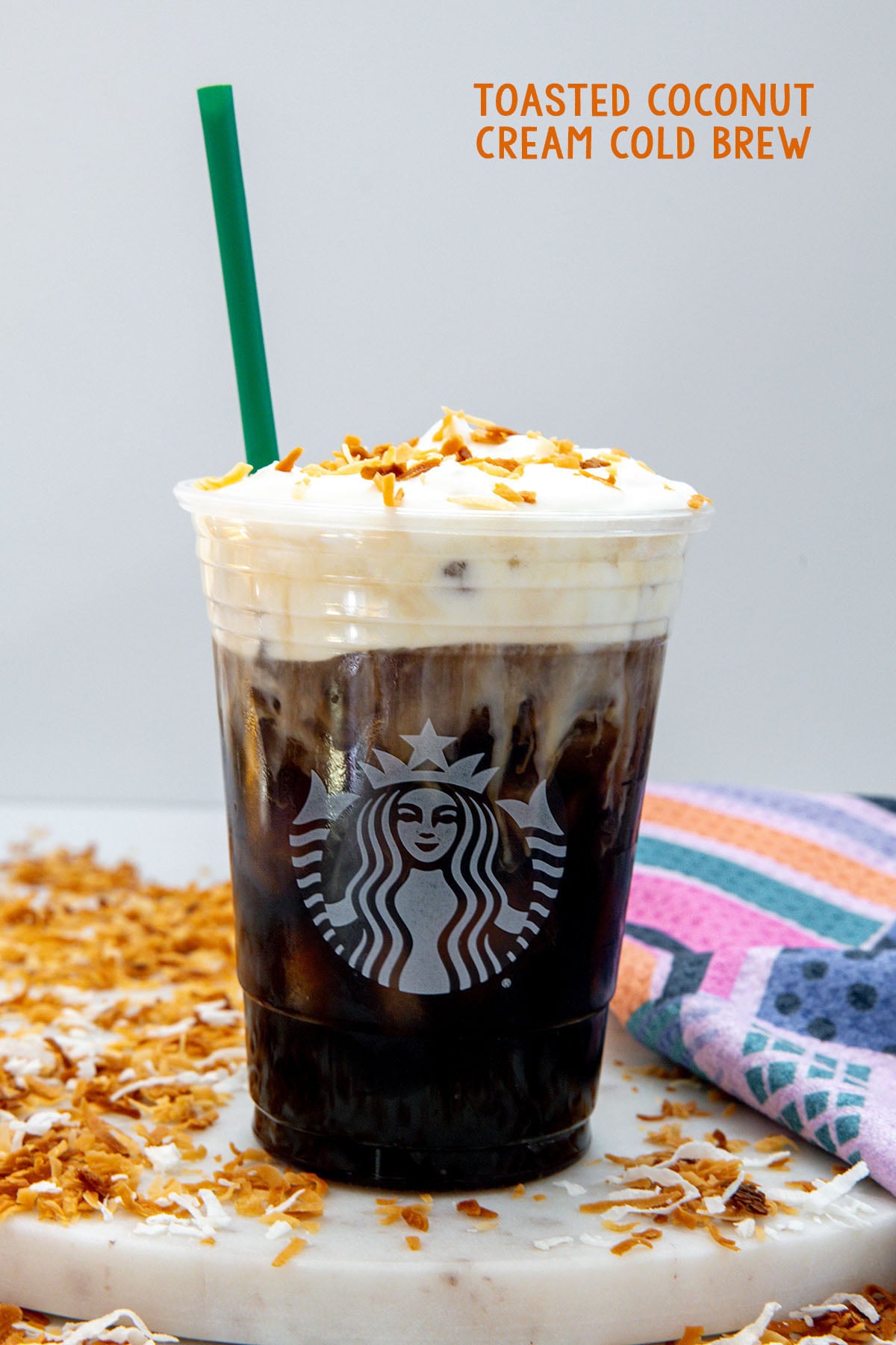 Head-on view of a Starbucks cup of cold brew topped with cold foam and toasted shredded coconut with coconut flakes all around and recipe title at top.