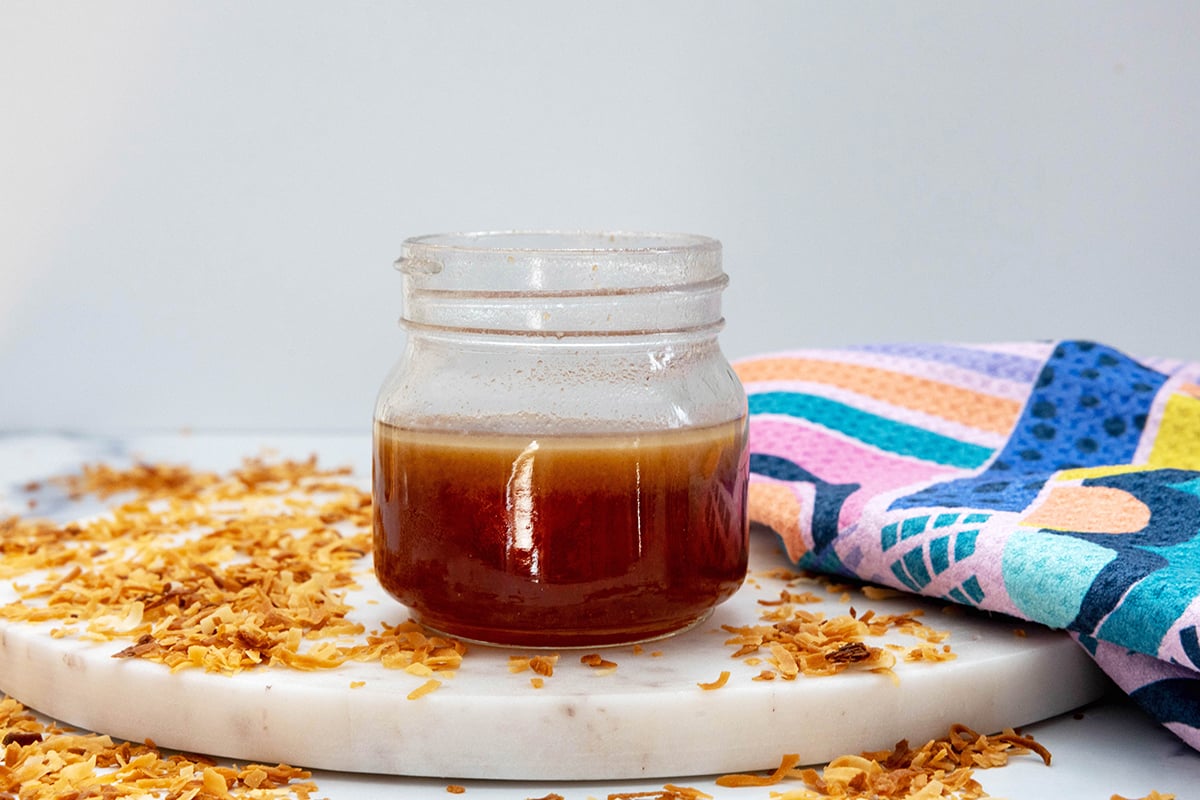 Head-on landscape view of a small jar of toasted coconut syrup with toasted coconut all around.