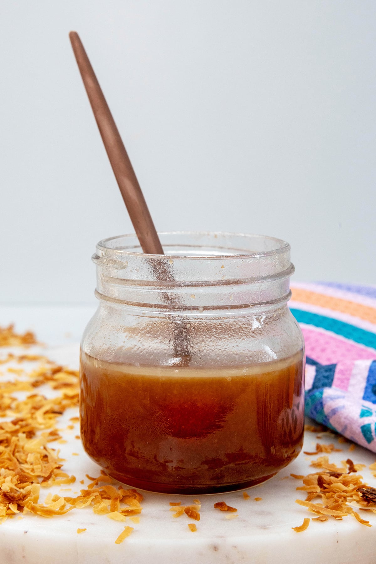 Head-on view of a small jar of toasted coconut syrup with spoon and toasted coconut all around.