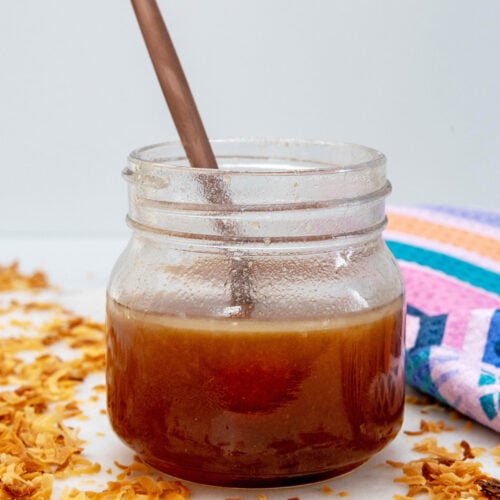 Head-on closeup view of a small jar of toasted coconut syrup with spoon and shredded coconut all around.