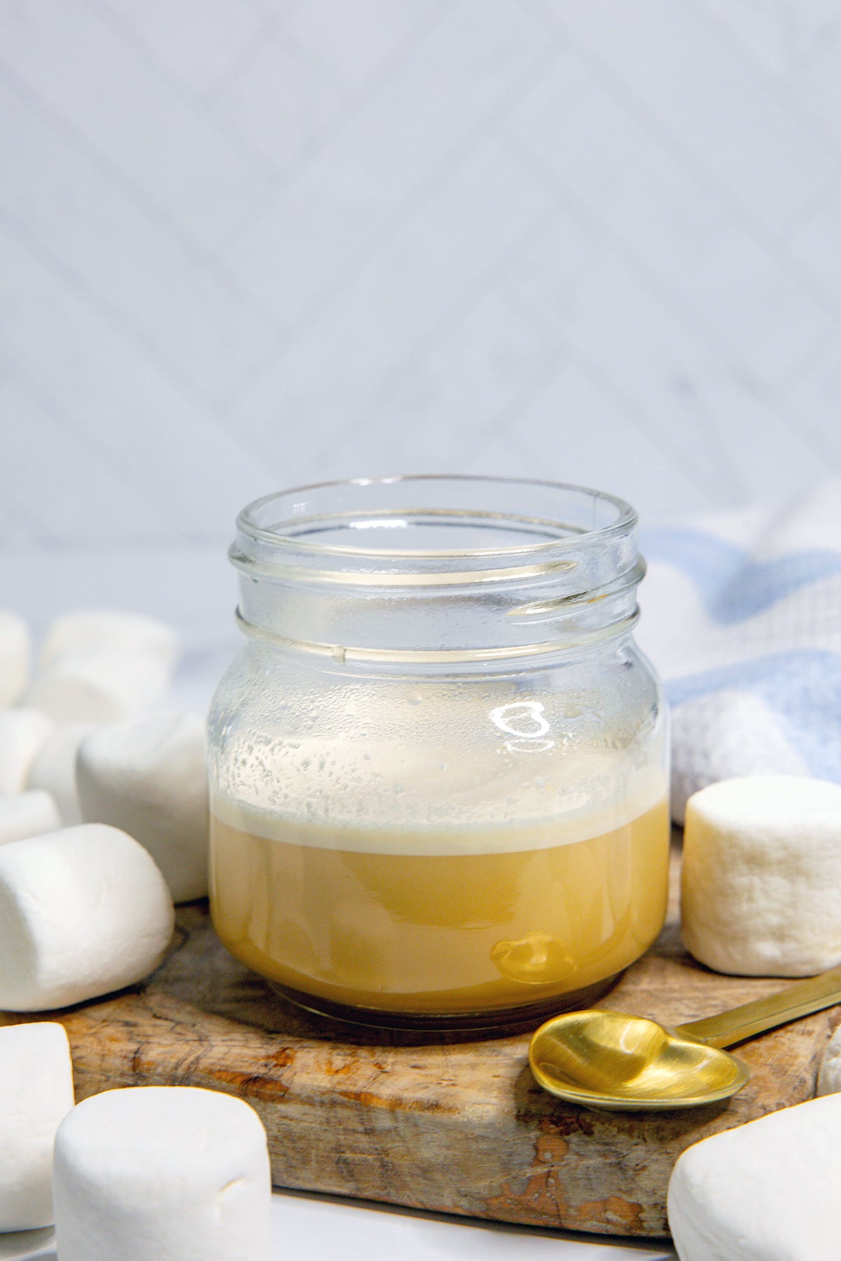 Head-on view of a small jar of toasted marshmallow syrup with marshmallows all around and heart-shaped spoon.