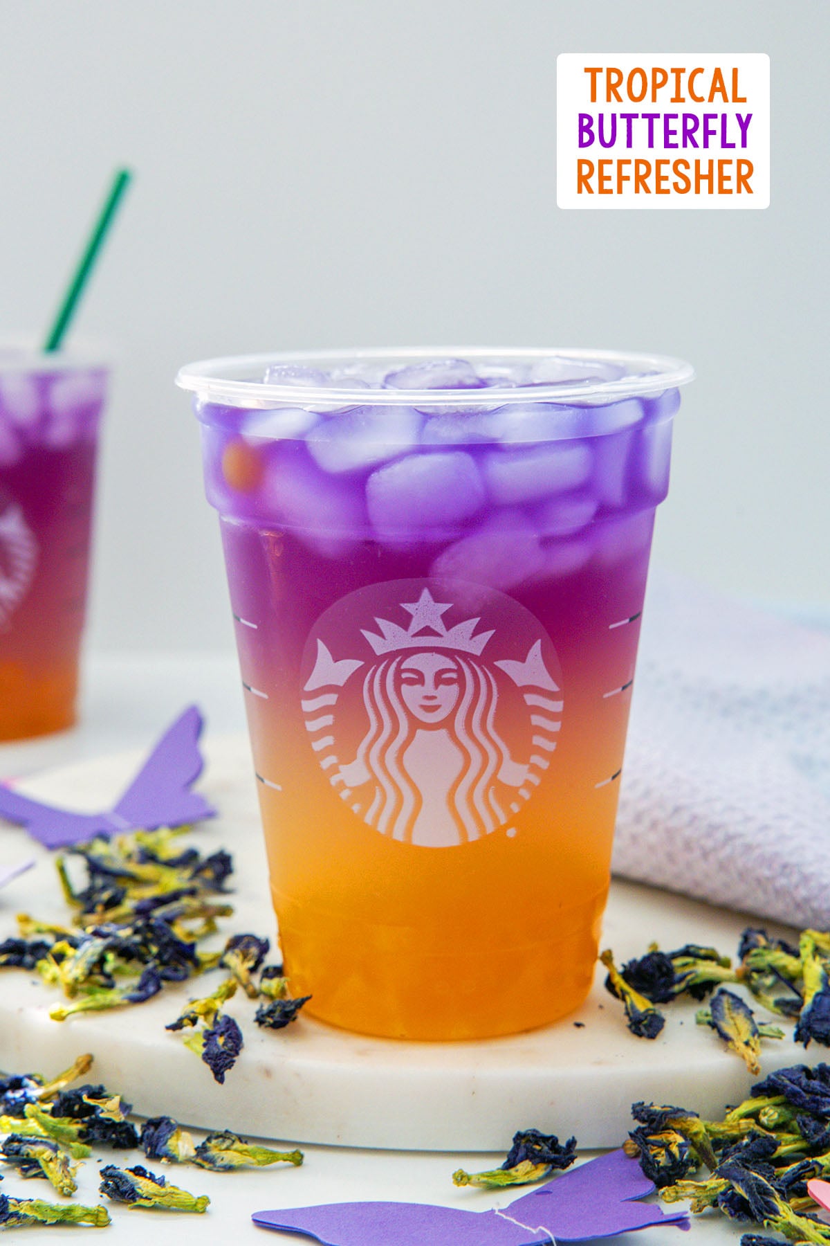 Head-on view of a Tropical Butterfly Refresher in a Starbucks cup with butterfly pea flowers all around and recipe title at top.