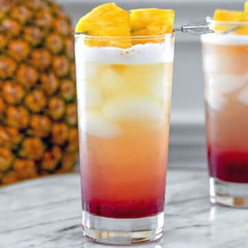Closeup view of a layered tropical gin sunrise drink with pineapple in background.