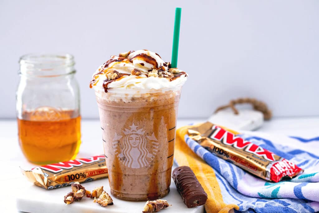 Landscape head-on view of a Twix Frappuccino with whipped cream and caramel sauce with Twix bars all around and jar of caramel syrup in background.