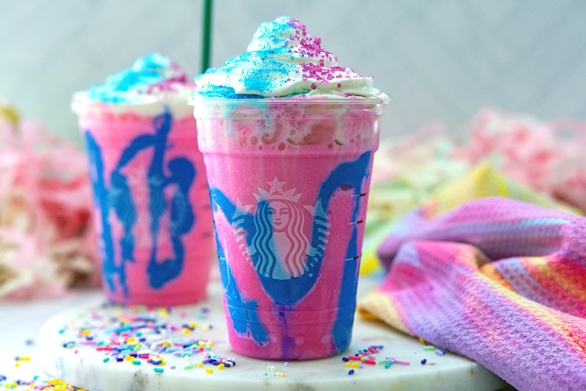 Head-on landscape view of two Unicorn Frappuccinos with whipped cream and blue and pink sugar.