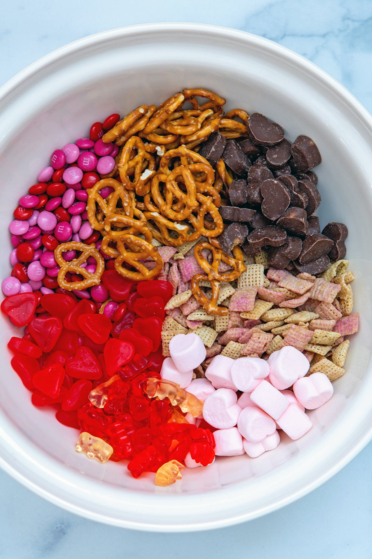 Big mixing bowl with Valentine snack mix ingredients separated, including heart marshmallows, heart peanut butter cups, heat gummies, M&Ms, pretzels, and Life cereal.