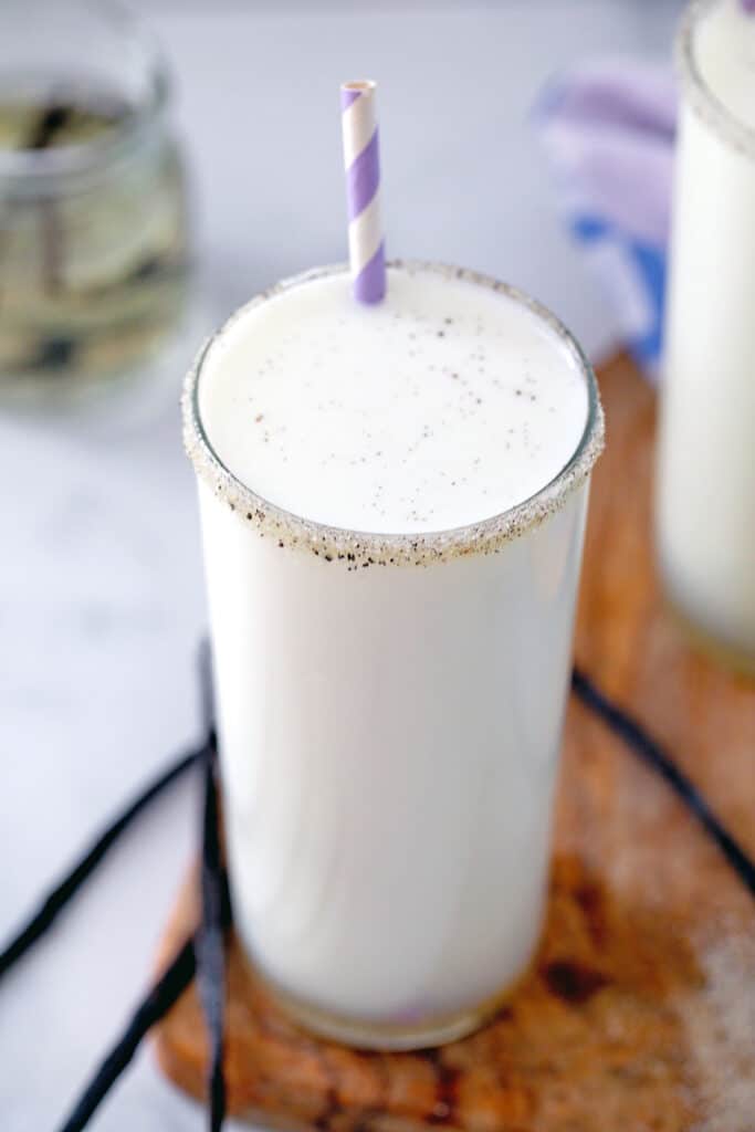 Overhead view of a glass of vanilla milk with purple and white straw and vanilla beans all around.