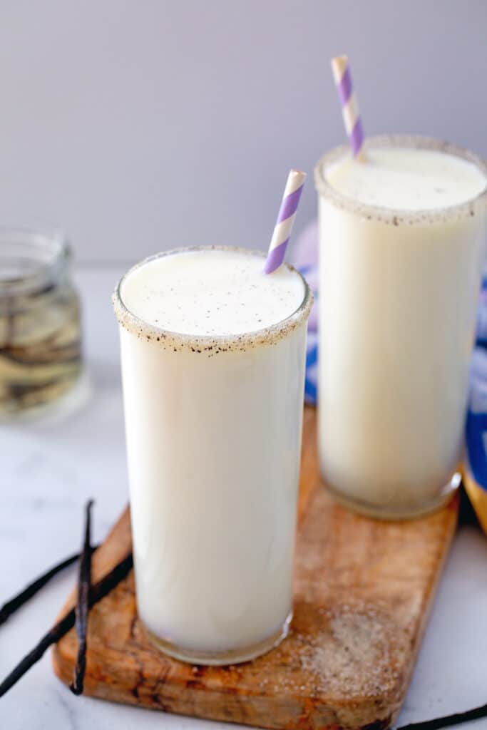 Two glasses of vanilla milk on a wooden board with vanilla beans all around.