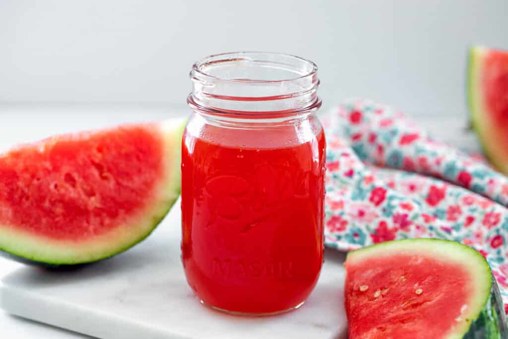 Landscape view of a mason jar of watermelon syrup with sliced watermelon in the background.