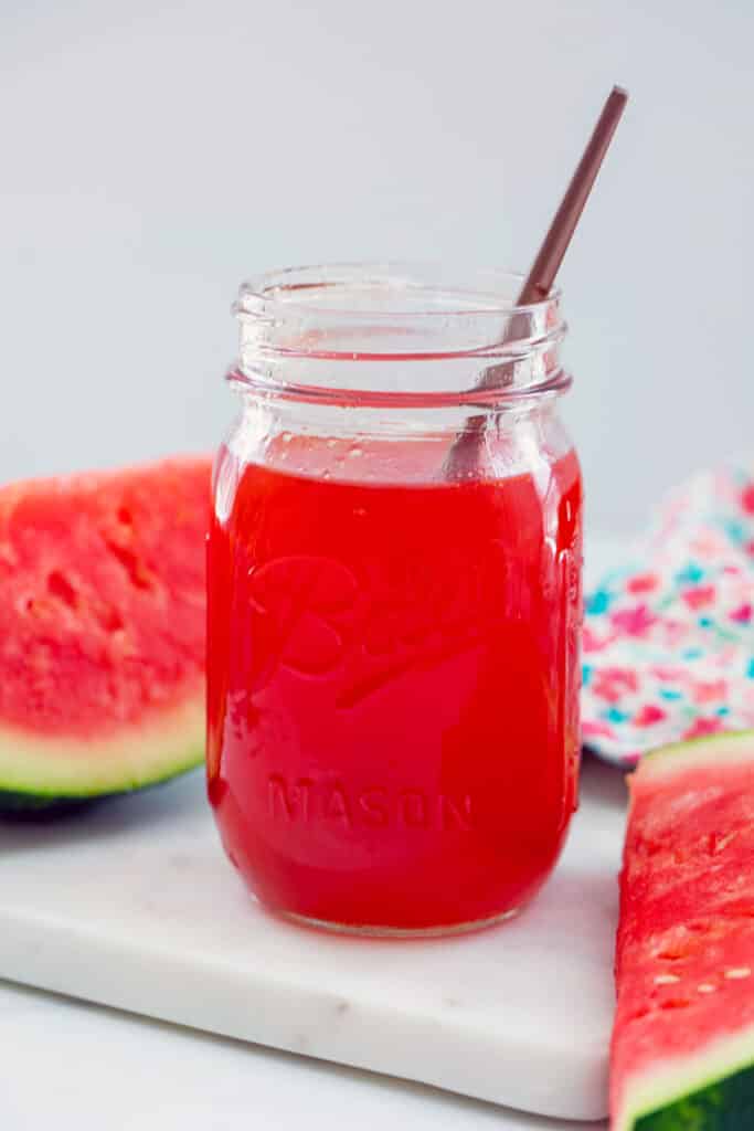 Mason jar of watermelon syrup with spoon in it with slices of watermelon in background.