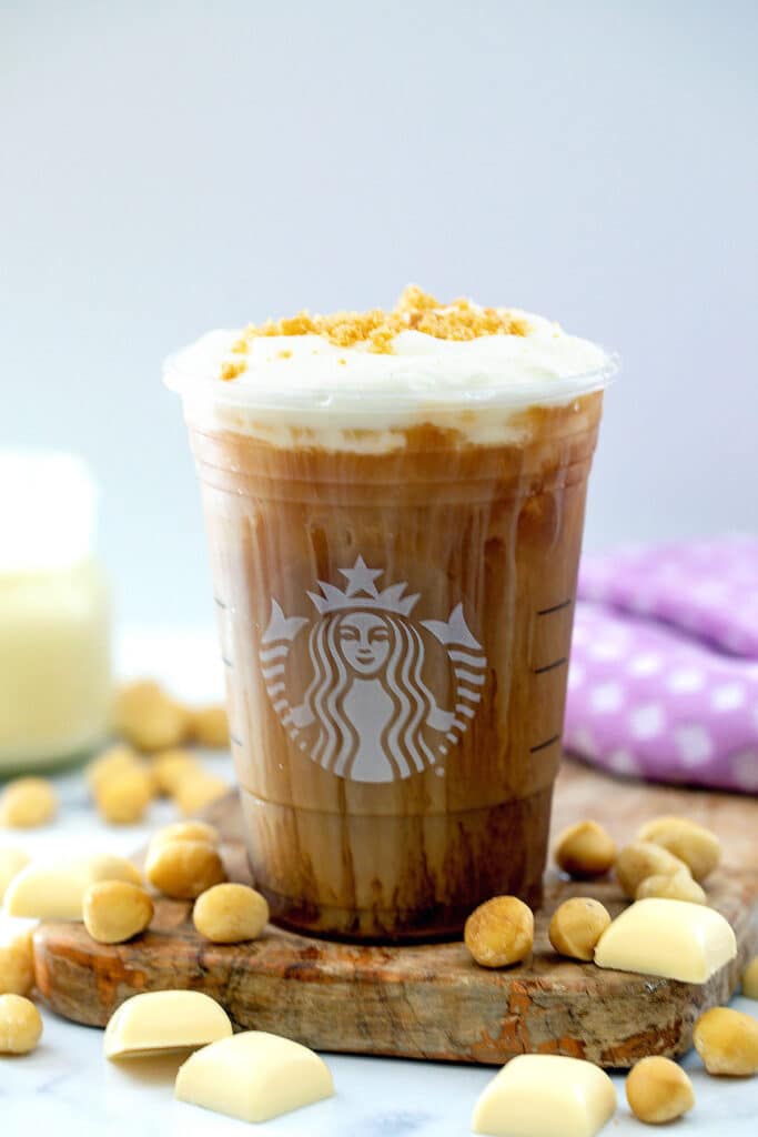 Head-on view of White Chocolate Macadamia Cream Cold Brew in Starbucks cup with nuts all around and small jar of white chocolate mocha in background.