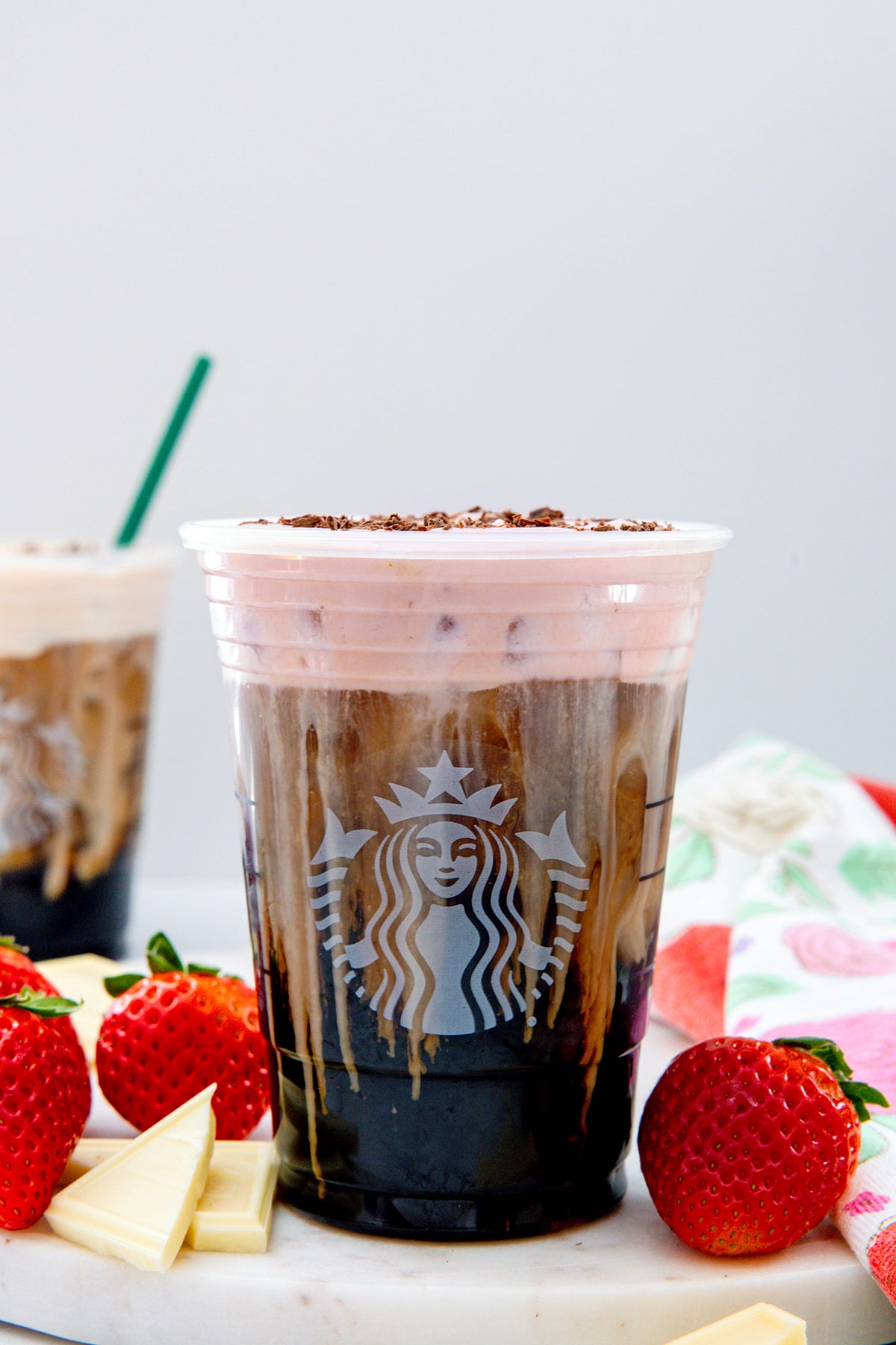 Head-on view of a White Chocolate Strawberry Cream Cold Brew in a Starbucks cup with second cup in background and strawberries and white chocolate all around.
