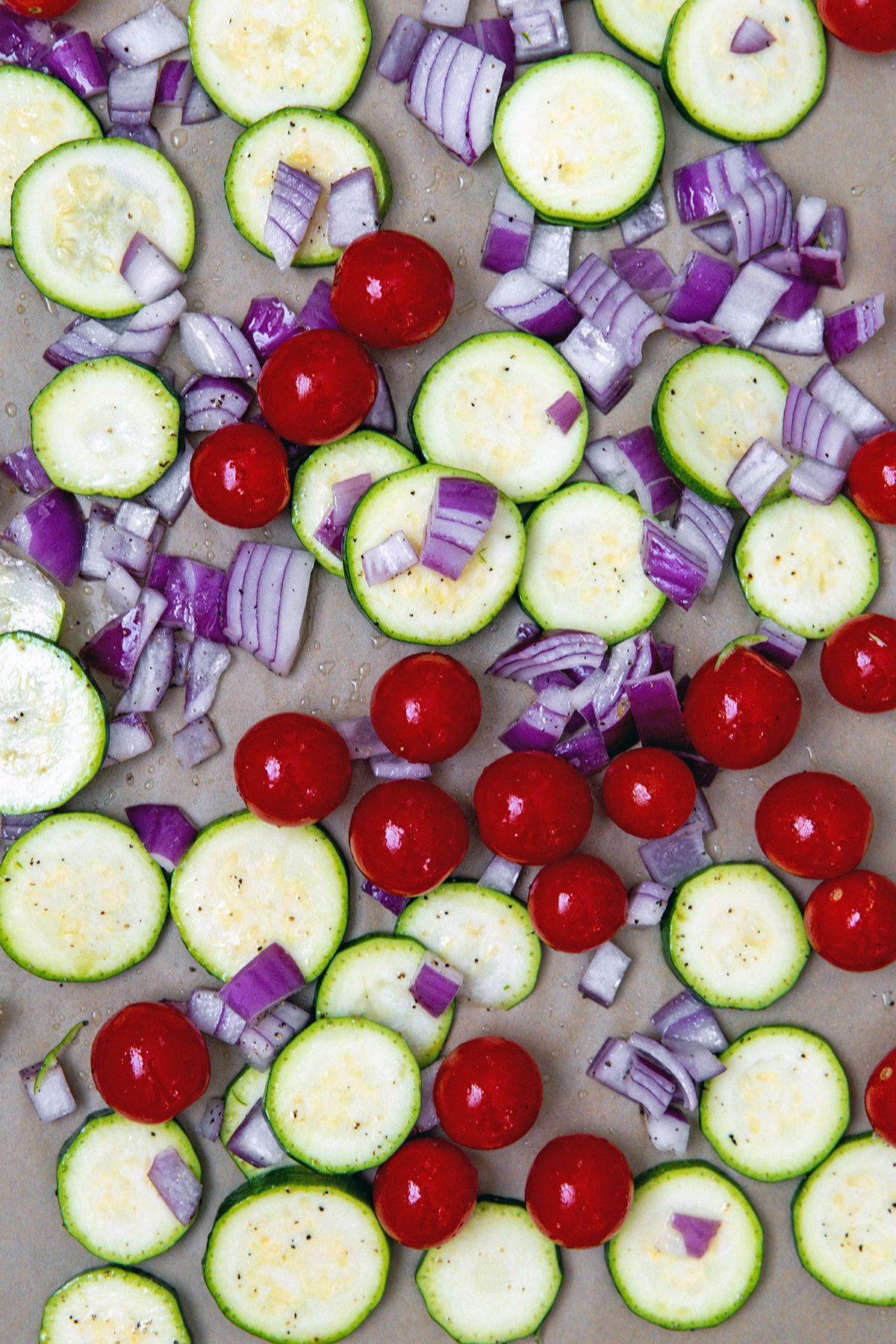 Overhead view of sliced zucchini, tomatoes, and red onion on parchment paper lined baking sheet.